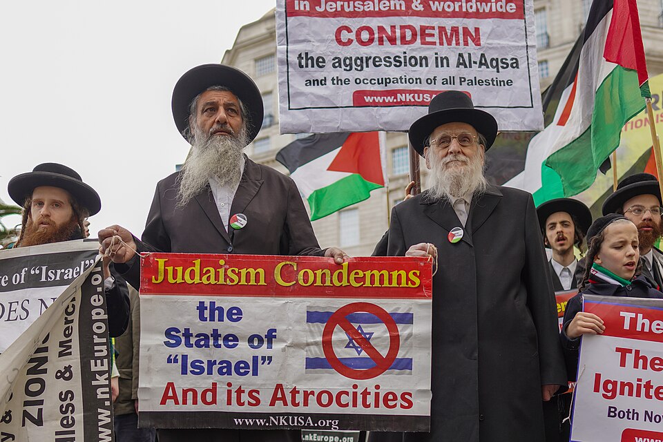 Anti-Zionist Haredi Jews at a protest holding a sign reading 'Judaism Condemns the State of Israel and its Atrocities' alongside Palestinian flags.